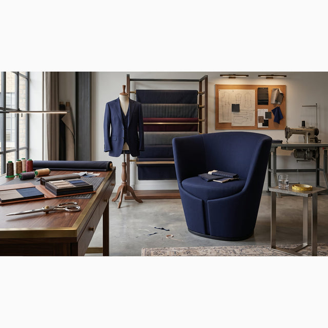 A navy blue Dapper Armchair in a tailor's workshop, paying homage to the inspiration behind its design. The room features a wooden work table with fabric swatches and scissors, a fabric rack with various coloured textiles, a mannequin wearing a navy blazer, and a sewing machine in the background.