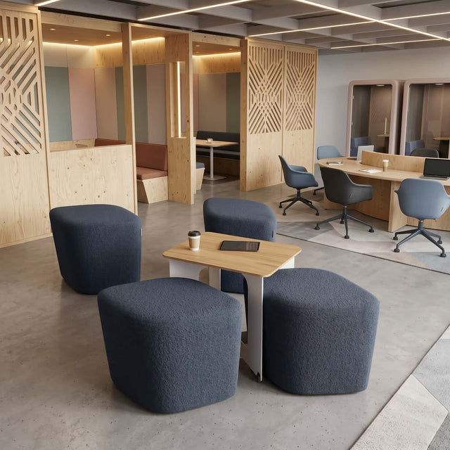 A communal space in a modern office features a Rhombic seating system with slate coloured poufs. One pouf remains neatly stowed away under the table, while the other 3 poufs have been pulled out for use.