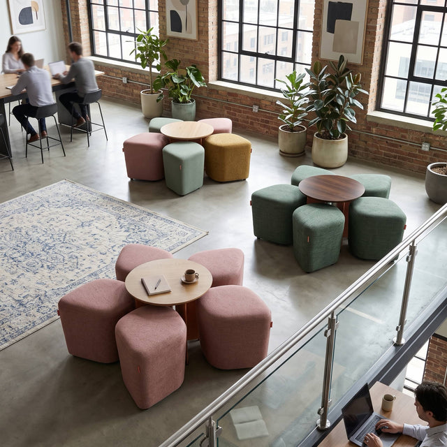 A modern loft-style breakout area in a workplace features three Blossom seating systems. Each Blossom consists of 5 upholstered poufs arranged around a central table, collectively forming the shape of a 5-petal flower.