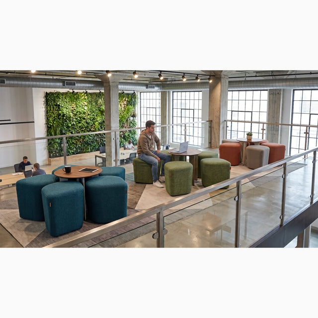 On the mezzanine of a modern office breakout area, three Blossom seating systems stand in a single line. A man sits at the middle station, working on a laptop placed atop its integrated table.
