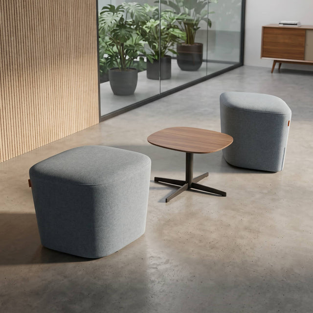 Two modern upholstered ottomans sit either side of a small wooden side table in a quiet corner of a bright, contemporary office.