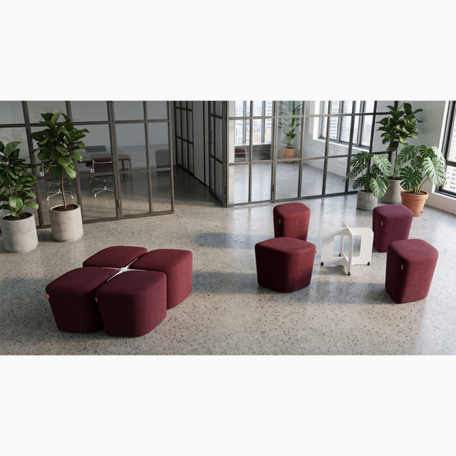 Two sets of the Rhombic seating system furnish a communal area in a workplace. The poufs are upholstered in maroon fabric and the docking bases are white.