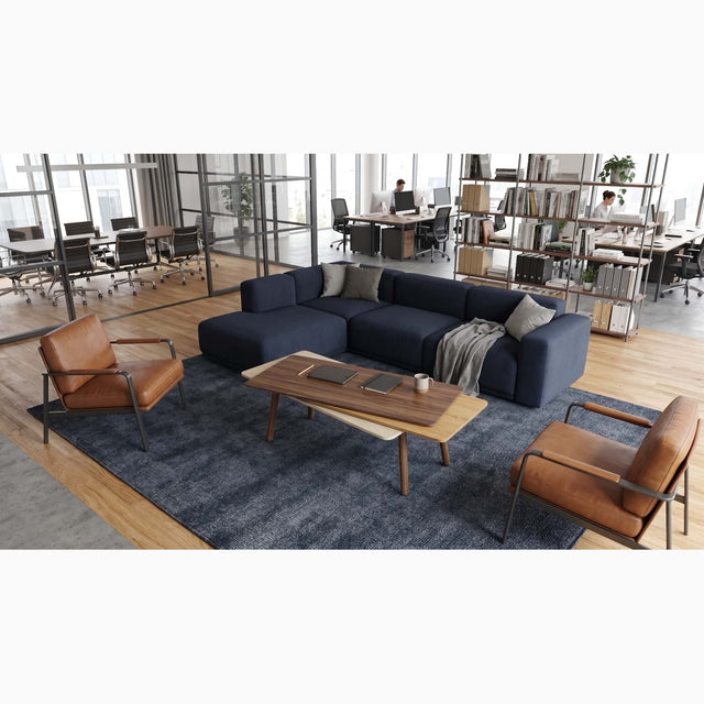 A Stacked Coffee table in wood features in an open plan office space. The table is surrounded by a blue sectional and 2 leather armchairs, and is made with a Walnut top surface, Oak middle and Ash bottom, along with Walnut legs.