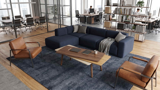 A Stacked Coffee table in wood features in an open plan office space. The table is surrounded by a blue sectional and 2 leather armchairs, and is made with a Walnut top surface, Oak middle and Ash bottom, along with Walnut legs.