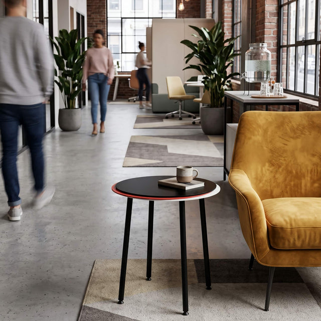 A black, red and white Stacked side table is  positioned next to a lounge chair in a thoroughfare of a busy office.