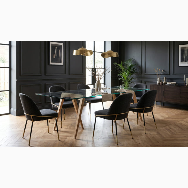 A modern dining room interior featuring a pair of Flex trestles being used with a glass tabletop to form a minimal, elegant dining table. It is surrounded by six black upholstered chairs with gold metal accents, and the room has dark panelled walls, a herringbone wooden floor, and gold pendant lights.
