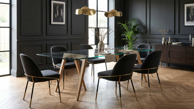 A modern dining room interior featuring a pair of Flex trestles being used with a glass tabletop to form a minimal, elegant dining table. It is surrounded by six black upholstered chairs with gold metal accents, and the room has dark panelled walls, a herringbone wooden floor, and gold pendant lights.