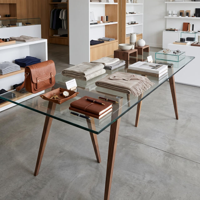 A pair of Flex trestle dining table legs in Walnut are paired with a glass tabletop to provide a merchandise display for clothing items in a styled boutique interior.