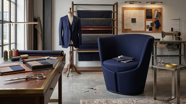 A navy blue Dapper Armchair in a tailor's workshop, paying homage to the inspiration behind its design. The room features a wooden work table with fabric swatches and scissors, a fabric rack with various coloured textiles, a mannequin wearing a navy blazer, and a sewing machine in the background.