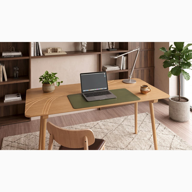 A Terrace Desk in Oak sits in a modern home office with wooden surfaces, warming brown tones and a beige rug. The desk is populated with a desk mat, laptop, mug, plant pot and articulated desk lamp.