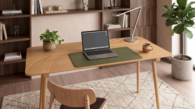 A Terrace Desk in Oak sits in a modern home office with wooden surfaces, warming brown tones and a beige rug. The desk is populated with a desk mat, laptop, mug, plant pot and articulated desk lamp.