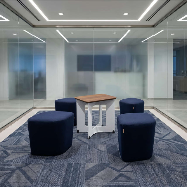 The Rhombic seating system is a glass-walled meeting room. Four blue poufs are scattered around the central table with a white frame and walnut tabletop.