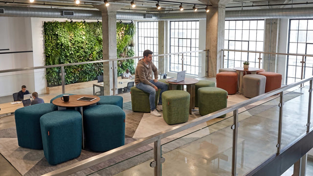 On the mezzanine of a modern office breakout area, three Blossom seating systems stand in a single line. A man sits at the middle station, working on a laptop placed atop its integrated table.
