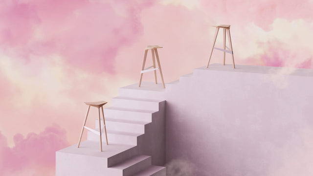 Three solid Oak Delta bar stools perched on a staircase high up in the clouds in an abstract sunset setting.
