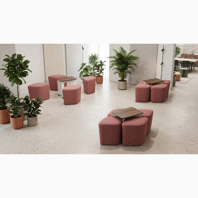 Three Rhombic seating systems with blush pink poufs and walnut tables adorn a co-working space in a busy office. One Rhombic system has its poufs pulled out from under its table ready for use, but the other two systems remain neatly squared away.