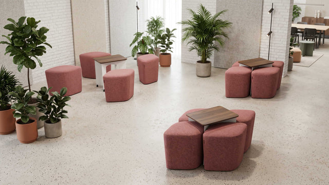 Three Rhombic seating systems with blush pink poufs and walnut tables adorn a co-working space in a busy office. One Rhombic system has its poufs pulled out from under its table ready for use, but the other two systems remain neatly squared away.