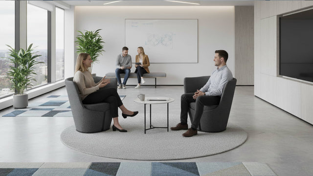Two people having a conversation whilst sat on grey Dapper armchairs in a communal office space.