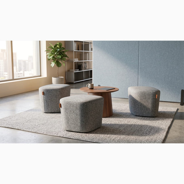 Three grey upholstered Petal poufs arranged around a small round wooden coffee table in a communal space of a bright and modern workspace.