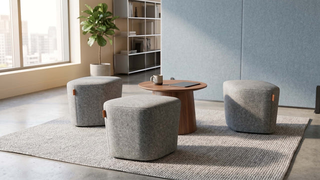 Three grey upholstered Petal poufs arranged around a small round wooden coffee table in a communal space of a bright and modern workspace.