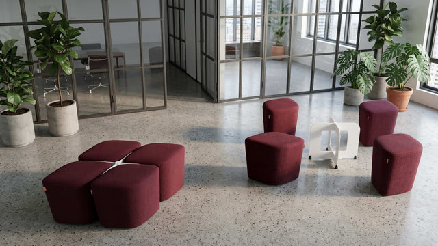 Two sets of the Rhombic seating system furnish a communal area in a workplace. The poufs are upholstered in maroon fabric and the docking bases are white.