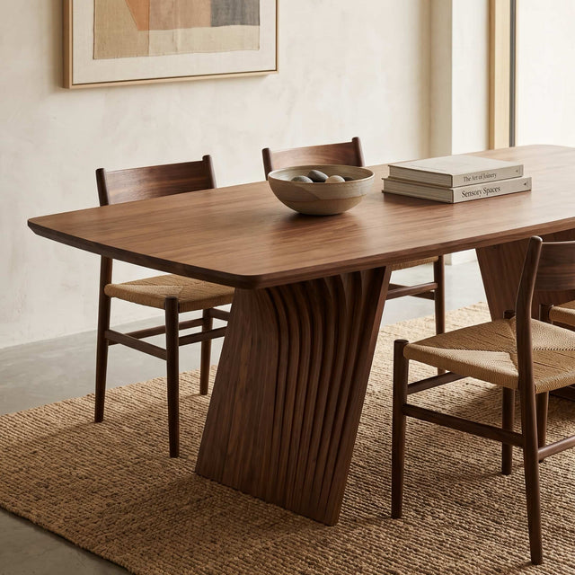 Close up of the legs of a Veer dining table in Walnut. The table features in a dining space along with wooden chairs, a rattan rug and contemporary wall art.